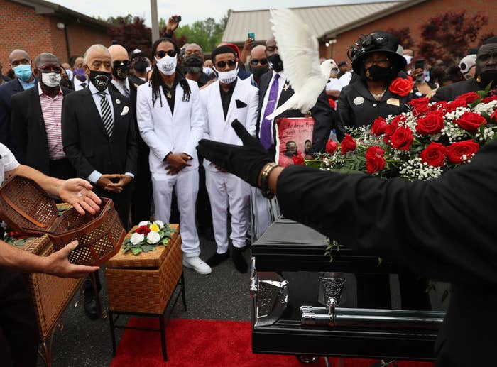Black men wearing suits and face masks stand around a casket at a funeral service