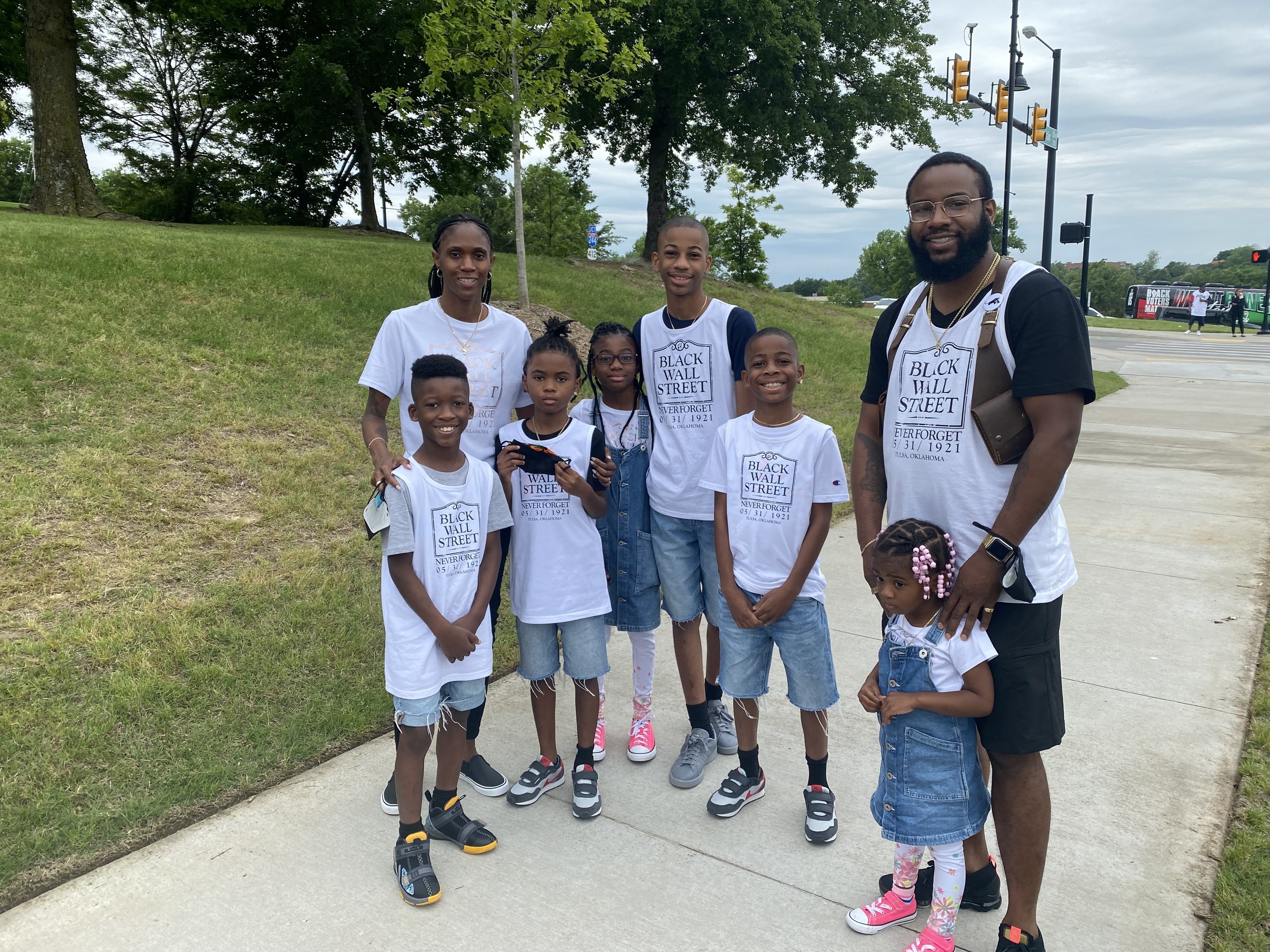 A family, including children, stands on the street, wearing &quot;Black Wall Street Never Forget 5/31/1921&quot; T-shirts