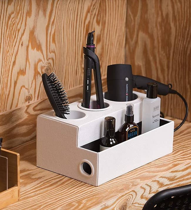 A white two tiered organizer with spots for a hairbrush, straightener, blowdryer, and hair products 