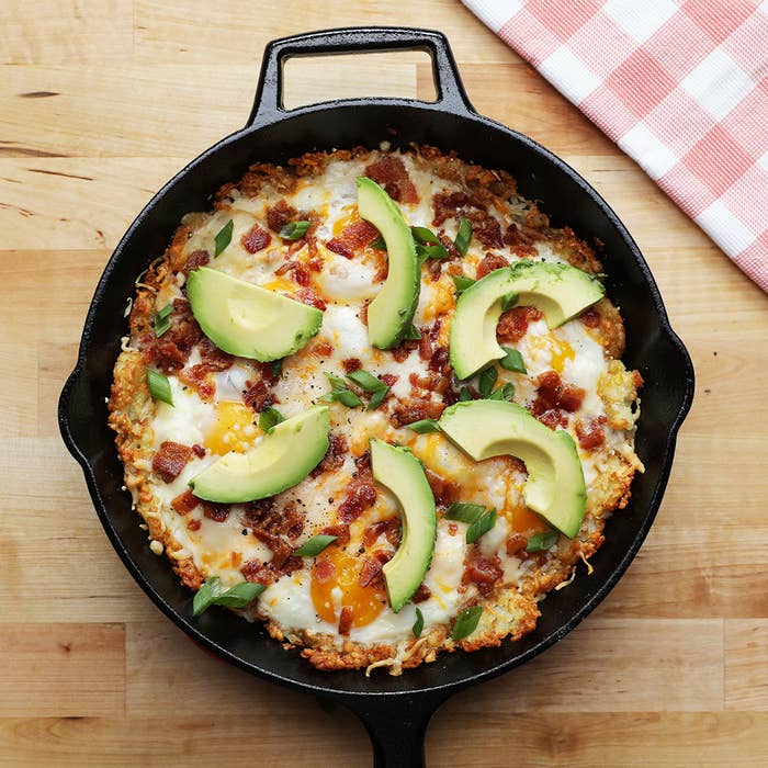 A skillet tater tot "pizza" topped with fried eggs, bacon bits, and avocado.