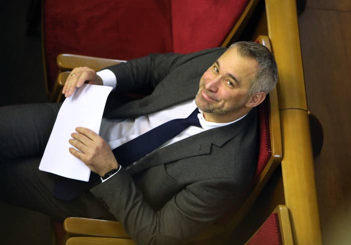 An aerial view of a man wearing a suit, sitting in a row of seats, holding a paper, looking up to the camera, and smiling