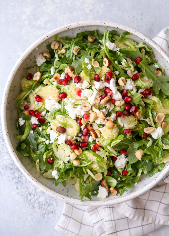 A bowl of arugula and Brussels sprouts salad topped with feta, hazelnuts, and pomegranate seeds. 