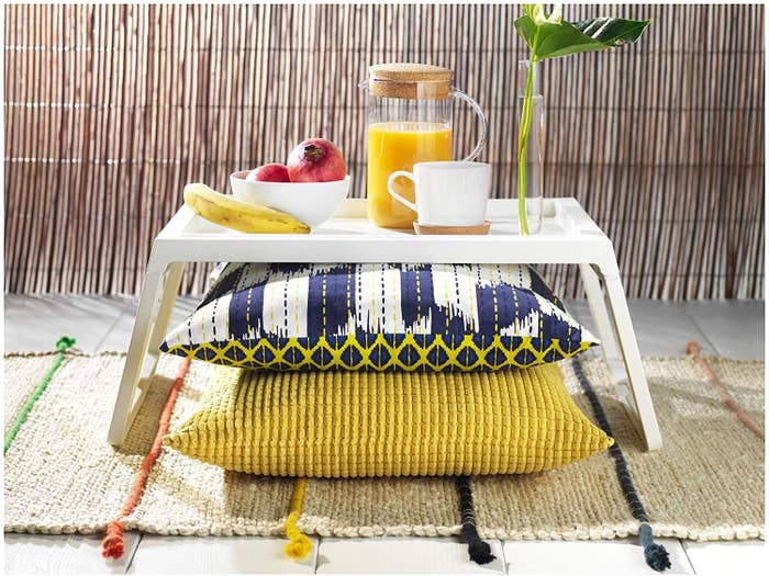 A foldable table with pillows under it. A cup of coffee, pitcher of juice, banana, and few pomegranates are kept on the table.