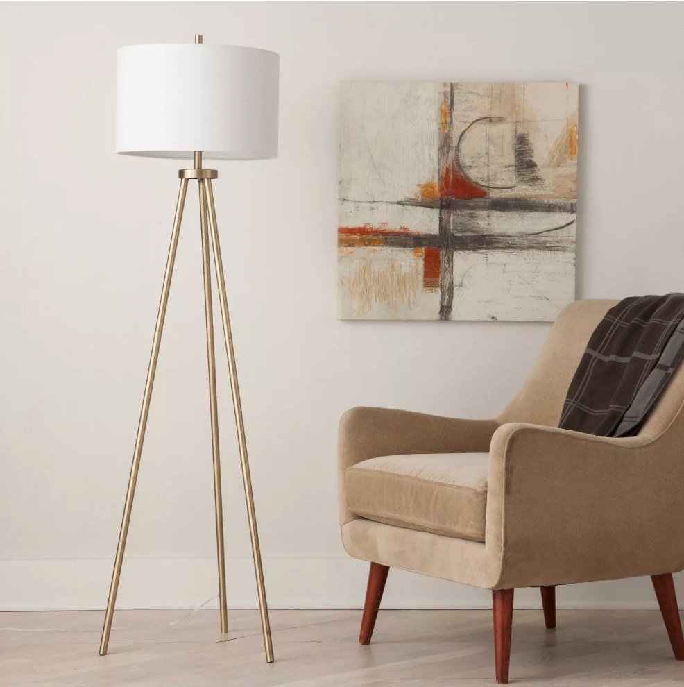 A sleek, brass, tripod floor lamp with a white drum displayed next to an accent chair