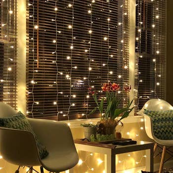 Glowing string light curtain hangs above living room window in front of small table and chairs 