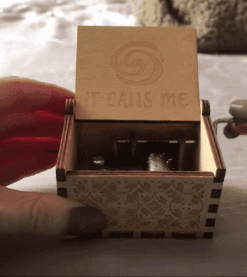 reviewer cranking up the wooden music box using the handle on the side. The box has a lid which is lifted up and has the heart of  Te Fiti engraved into it plus the words, &quot;It calls me.&quot;