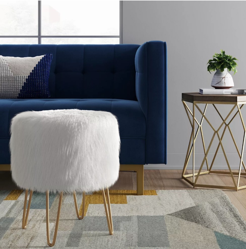 A white, faux fur ottoman on gold legs displayed in a living room 