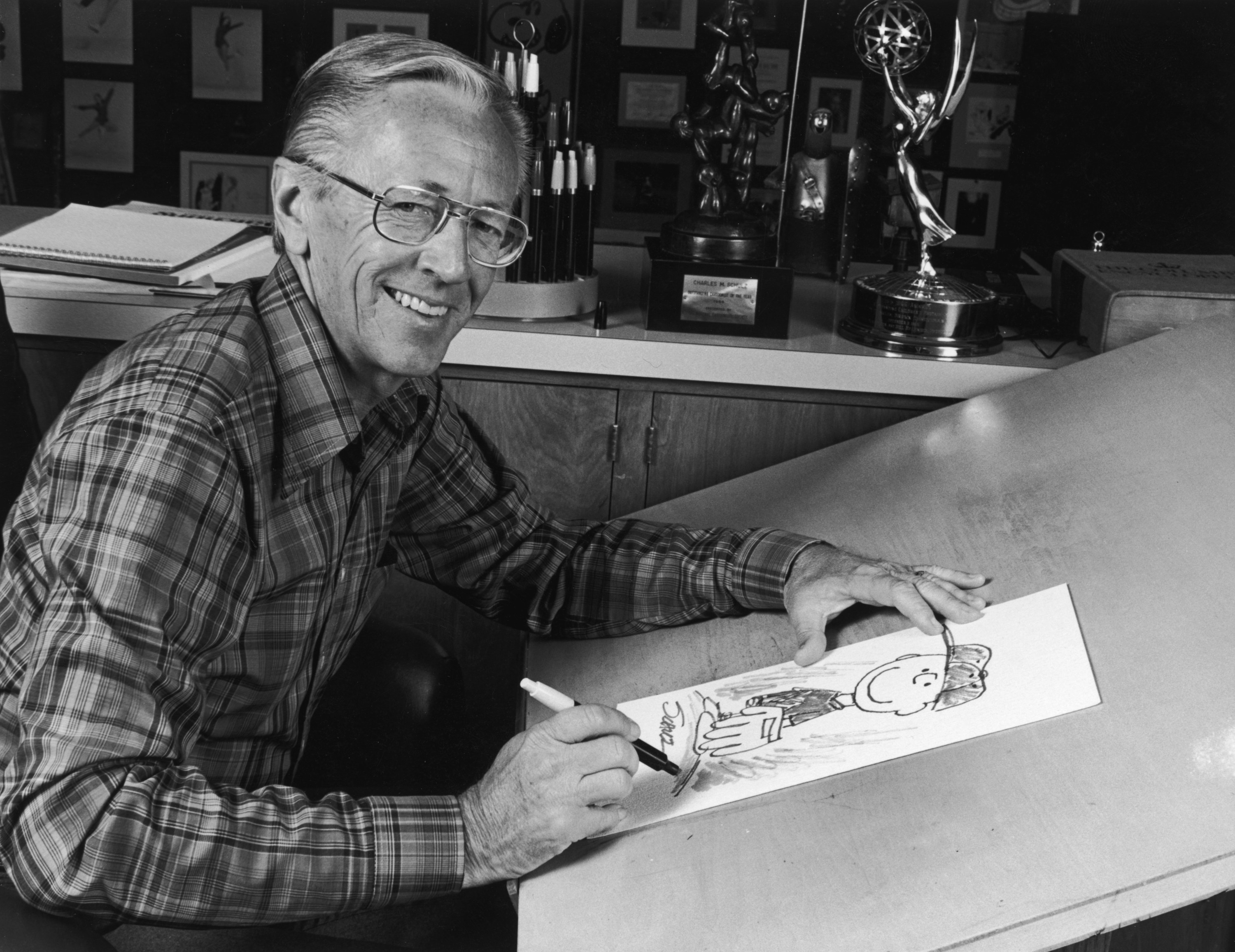 Black-and-white photo of Charles Schulz at a table drawing a sketch of Peanuts character Charlie Brown