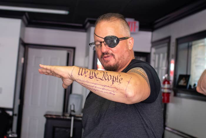 A man with an eyepatch holds up his forearm, which has a tattoo that reads &quot;We the people&quot;