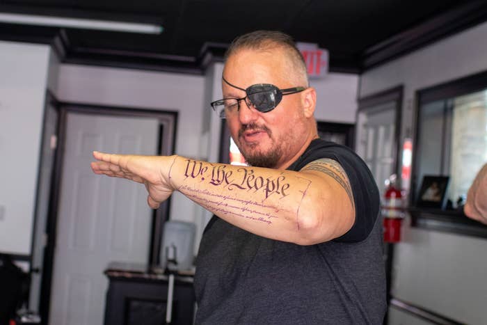 A man with an eyepatch holds up his forearm, which has a tattoo that reads "We the people"