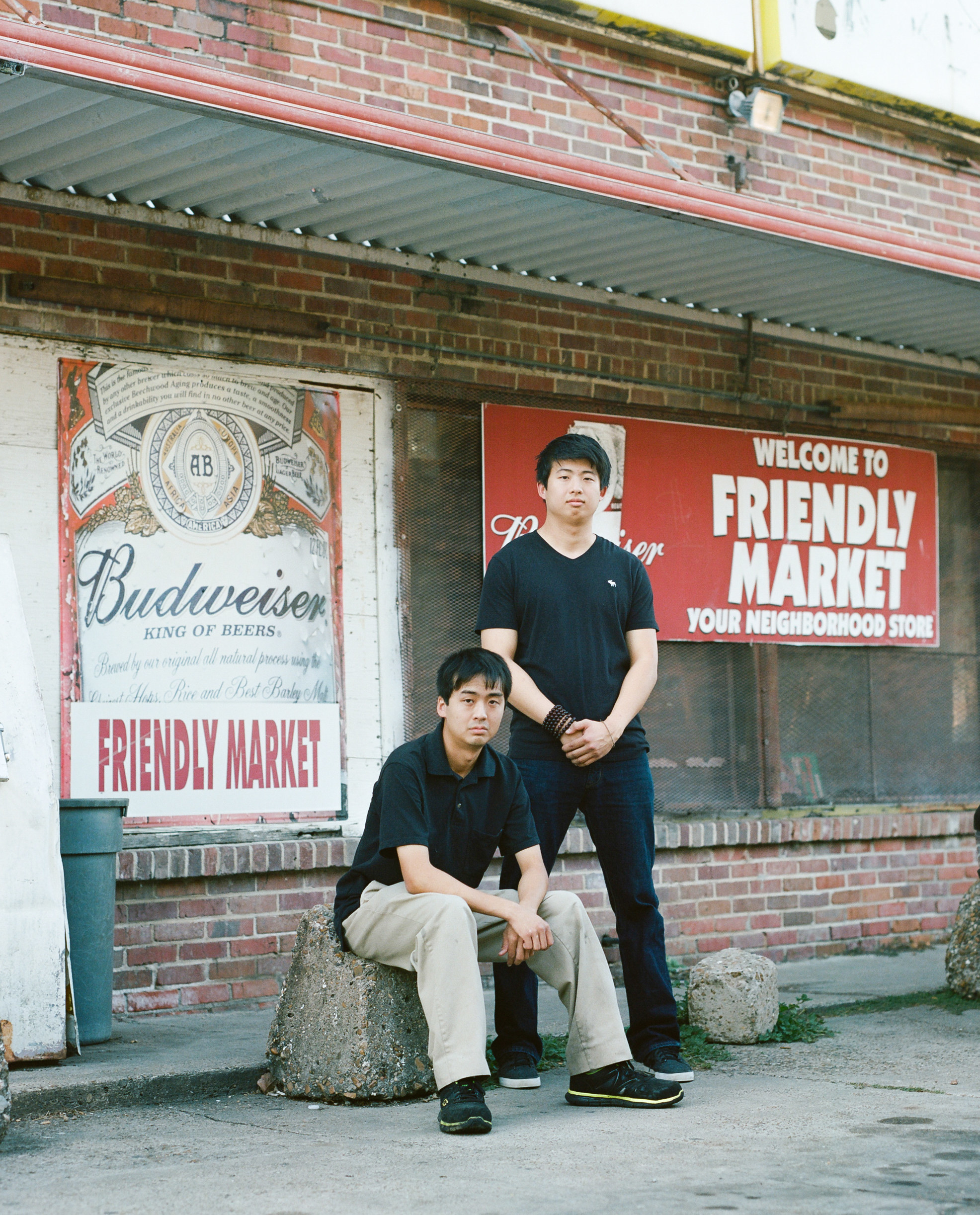 Two men sit in front of a small store with Budweiser signs and a sign saying Your Friendly Neighborhood Market