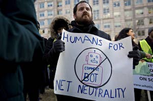 A demonstrator holds a poster that says "Humans For Net Neutrality." The poster also contains a drawing of a robot, crossed out and with "Comment Bot" written on its torso.