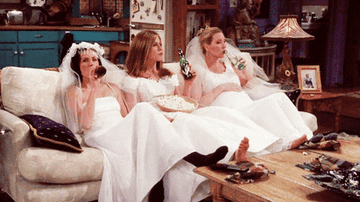Monica, Rachel, and Phoebe sit on the couch drinking beer and eating popcorn while wearing wedding dresses