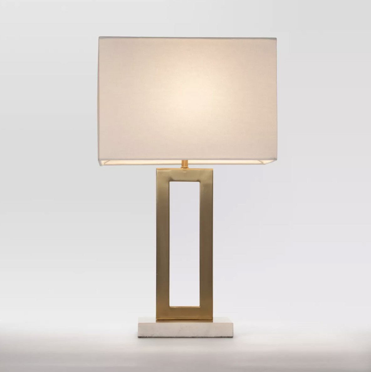 The lamp is on and has a white lampshade connected to a gold rectangular body that sits vertically on a white base 