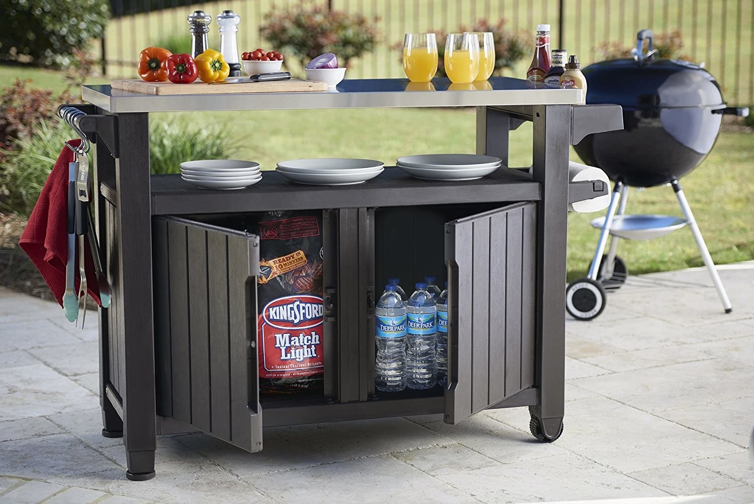 the unit with two plastic cabinet doors storing charcoal and water, a lower shelf stacked with plates, and a stainless countertop with a cutting board and drinks. On the side is a roll of paper towels, and the other holds grilling equipment
