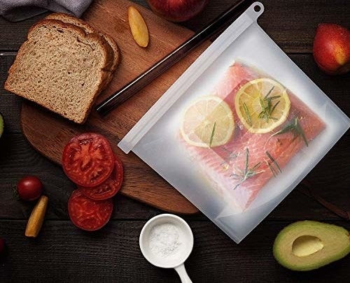 A fillet of salmon, lemon slices, and sprigs of herbs are kept inside the silicone bag. There are apples, peaches, slices of tomato, slices of bread, and half an avocado kept around the bag.