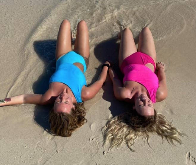 A reviewer showing herself and her sister laying on the beach wearing the matching swimsuit in blue and pink 