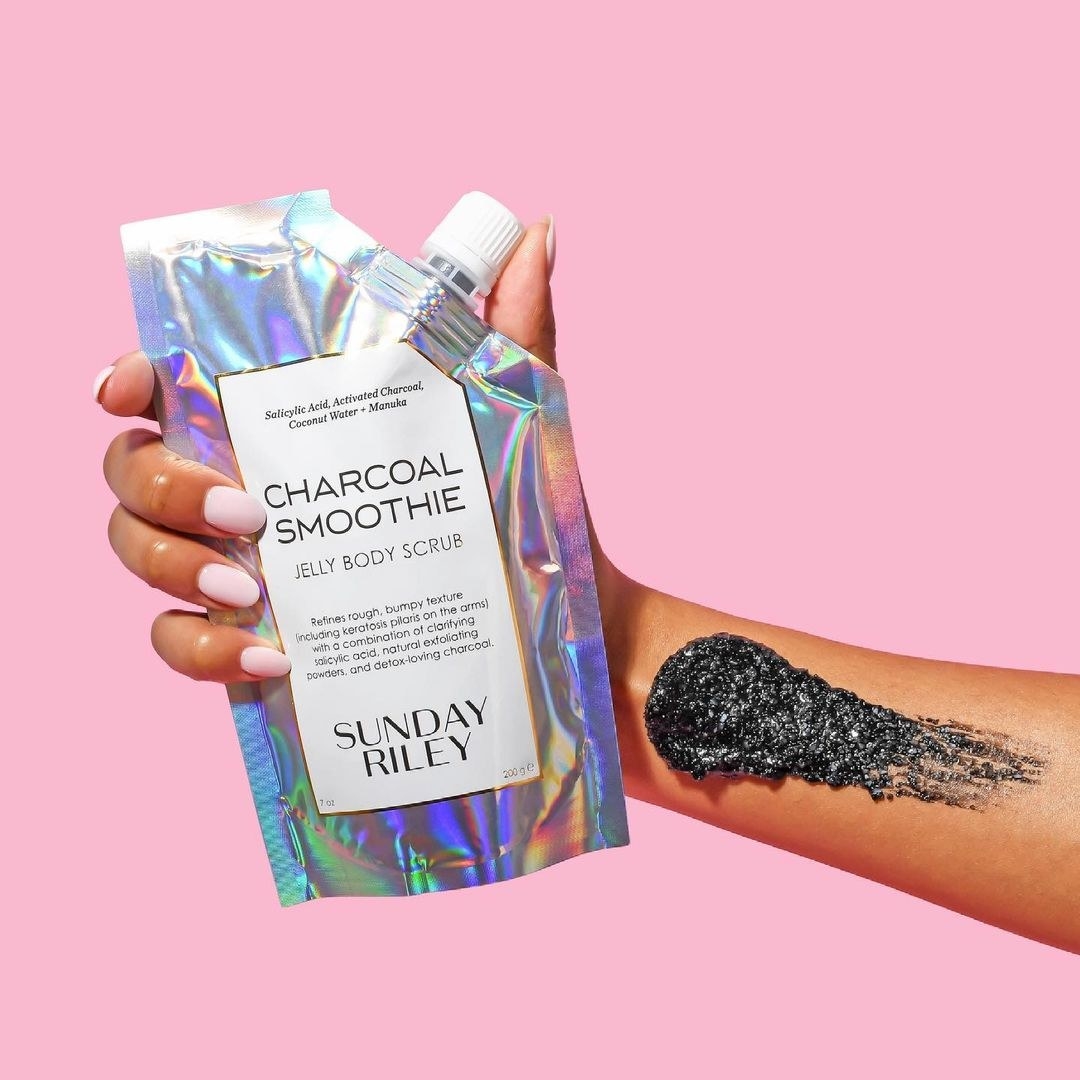A person holding the iridescent packaging with a smear of the charcoal scrub on their arm