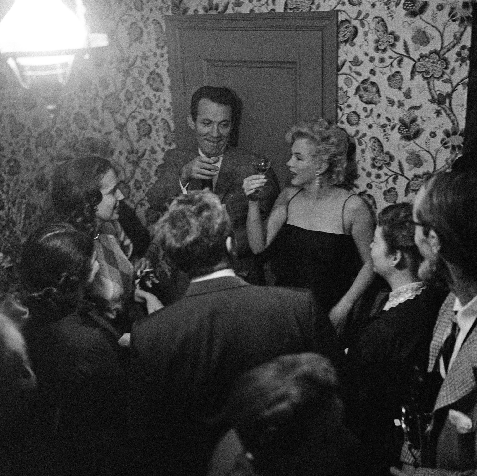 Marilyn Monroe holds up a small glass of alcohol, surrounded by people at a party