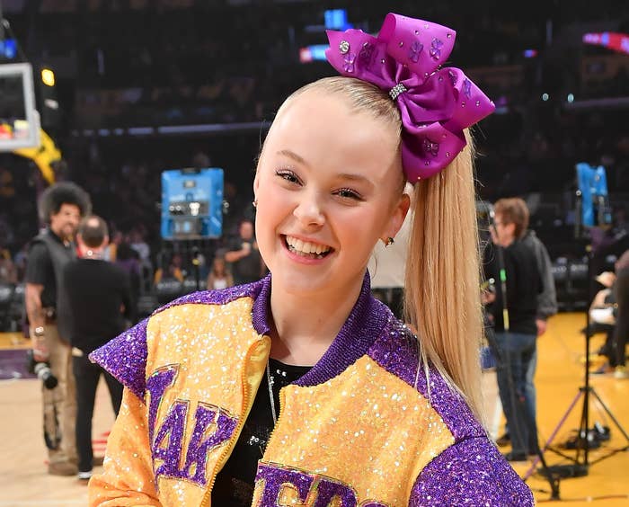 JoJo smiles while wearing a sparkly purple and yellow jacket and purple hair bow