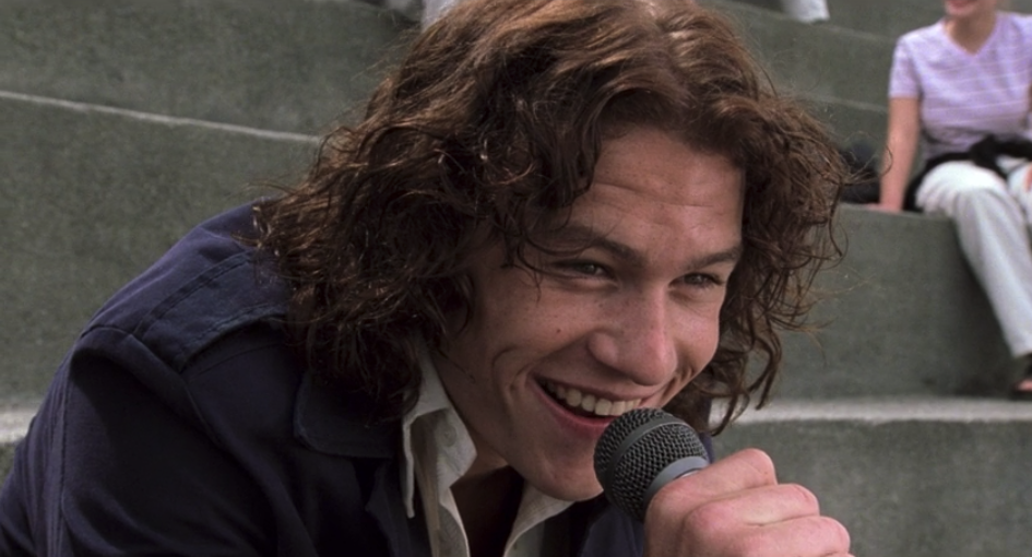 Heath Ledger in 10 Things I Hate About You; he is sitting on some steps while singing into a microphone 
