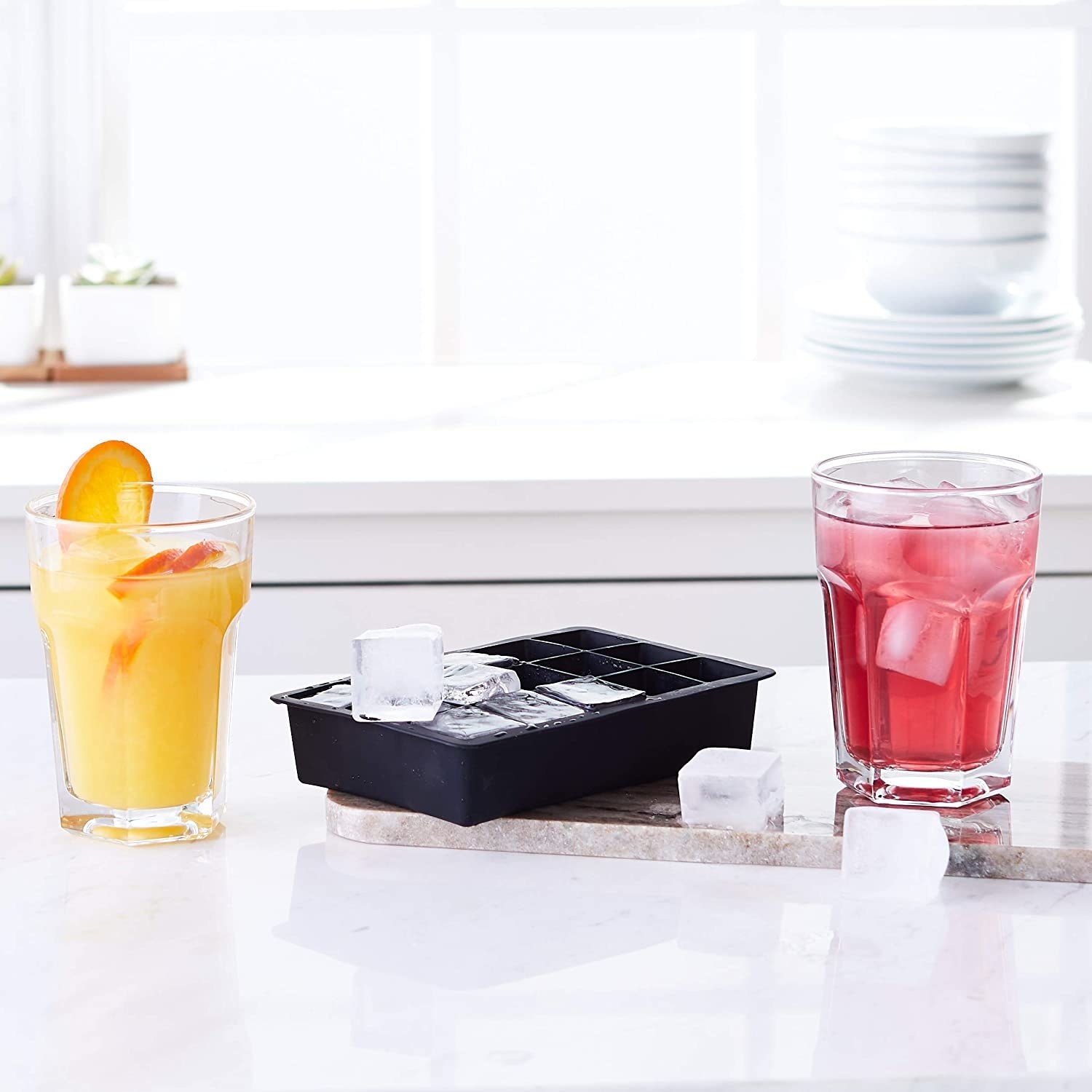 A black silicone ice cube tray with ice cubes surrounding it and two glasses with drinks in them