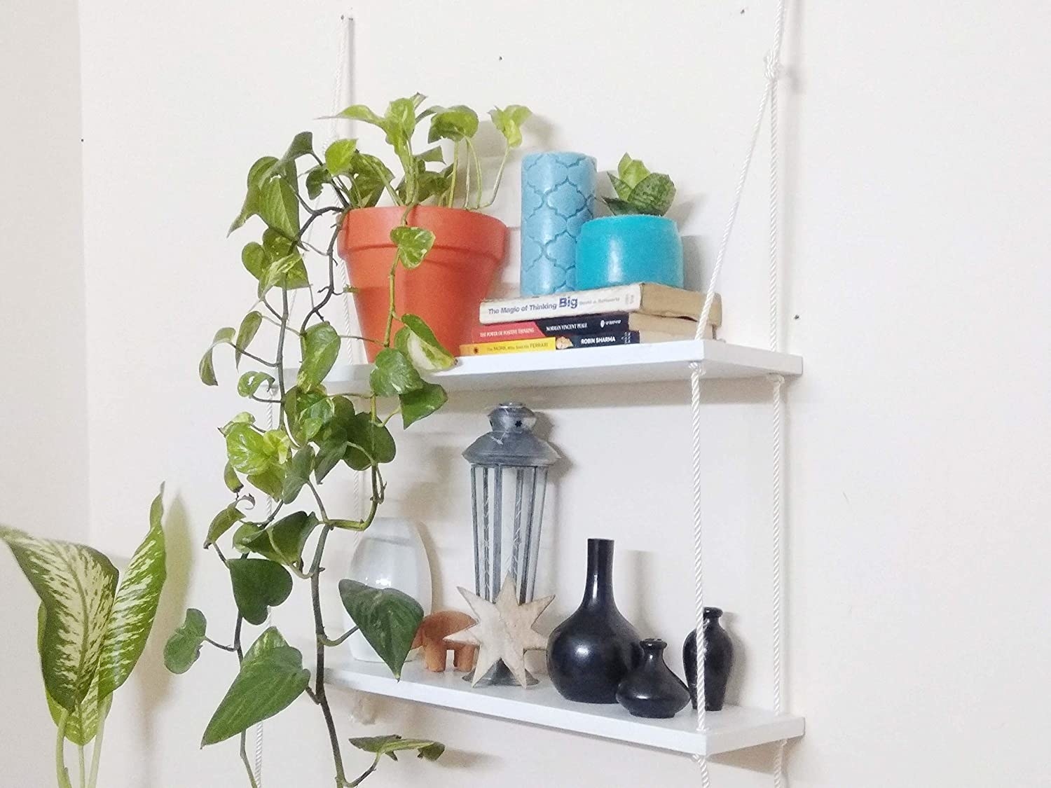 A two-tier shelf held together by sturdy ropes, with books, candles, and a plant on it.