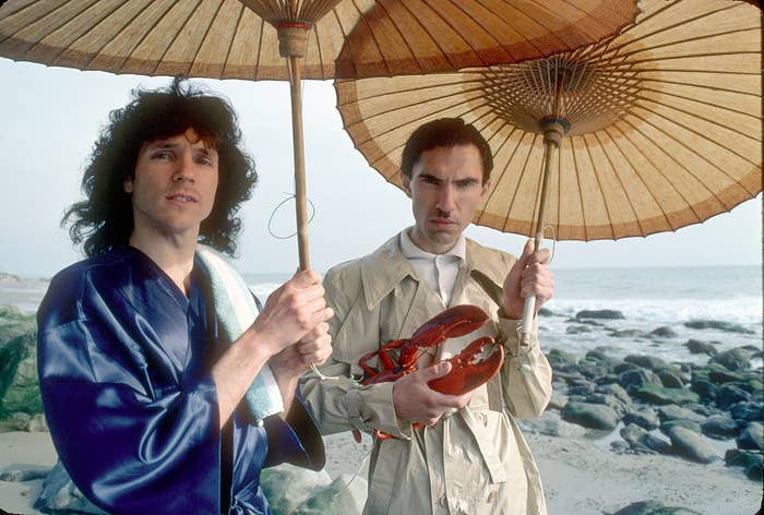 Russell wearing a silk robe and Ron wearing a trench coat, holding a fake lobster, both holding parasols, at the beach