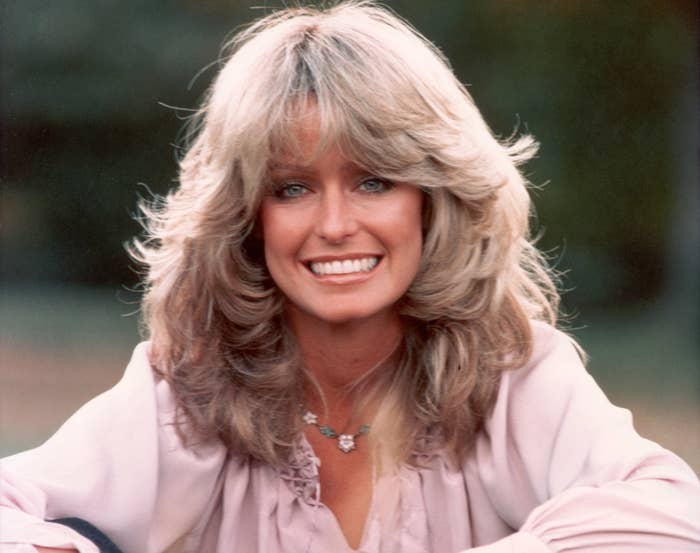 A young Farrah smiles while sitting outside in a while blouse