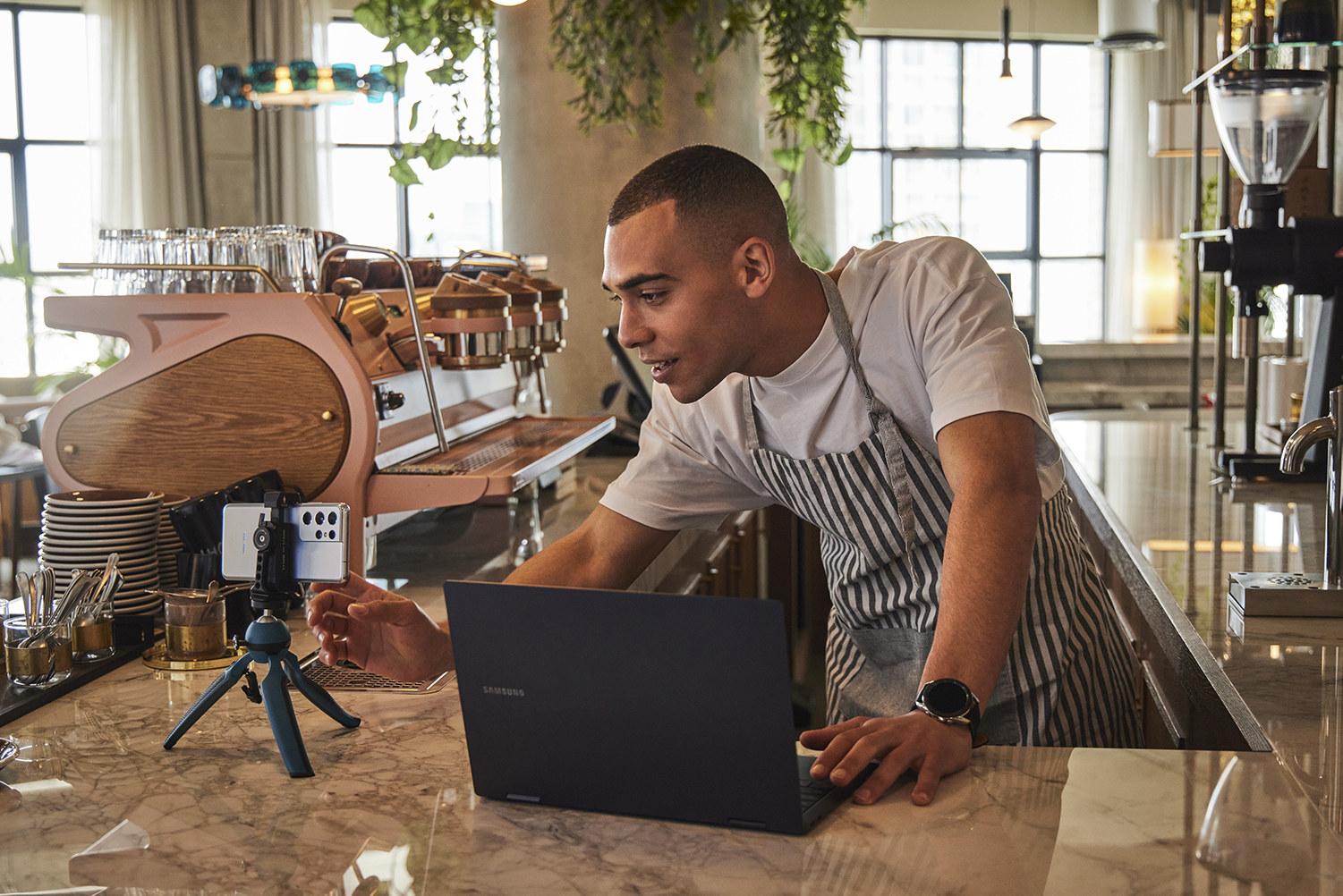 A man at a bar in a restaurant at a laptop touching his phone which is on a tripod.