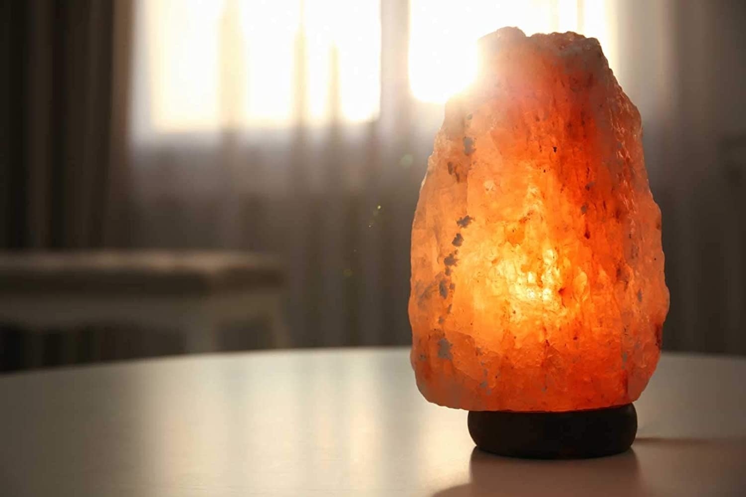 A Himalayan Rock salt lamp on a table with sunlight peeking through the curtains in the background.