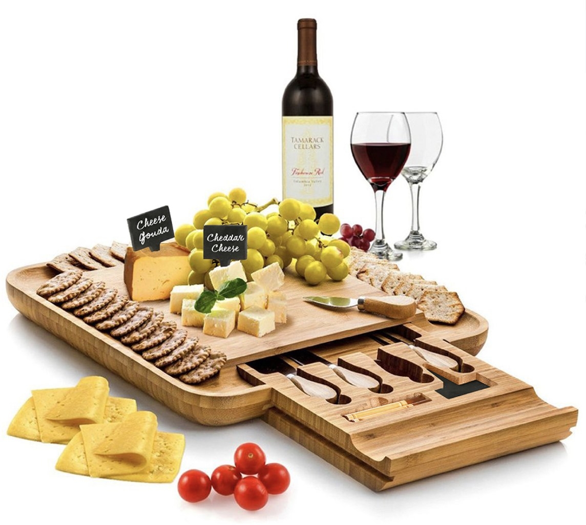 The tan board is holding cheeses, fruits and crackers and there is a pullout compartment with the utensils
