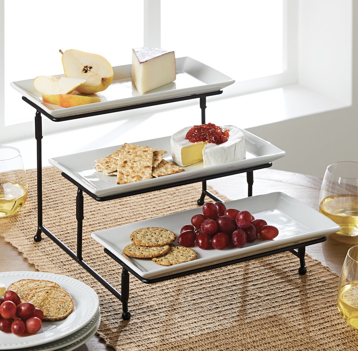 The white porcelain trays are staggered on a black metal frame and are holding fruit, cheese and crackers