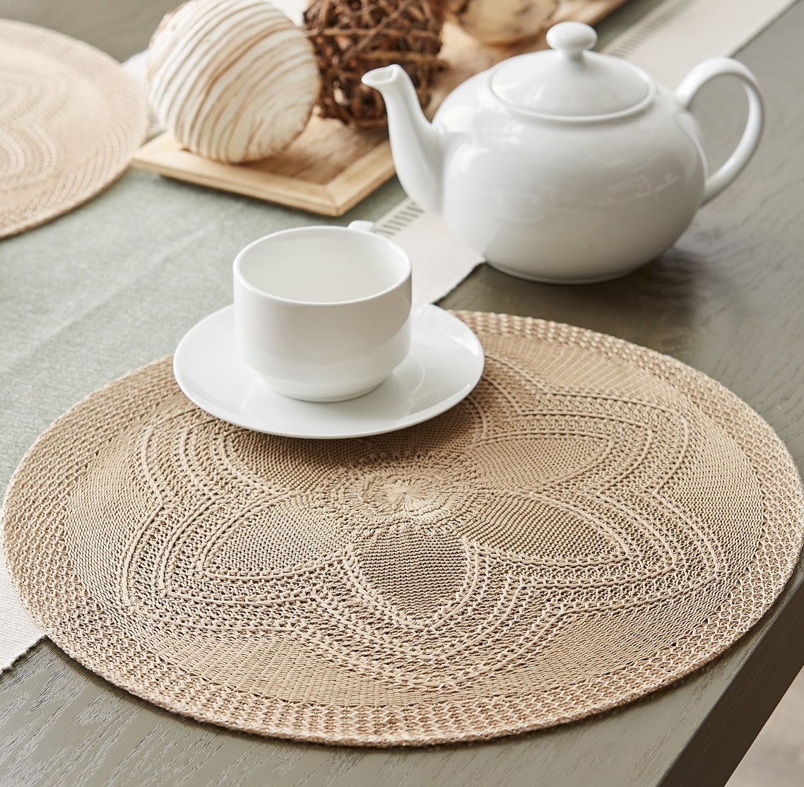 The tan floral stitch placemat is on a wooden table and surrounded by white porcelain decor