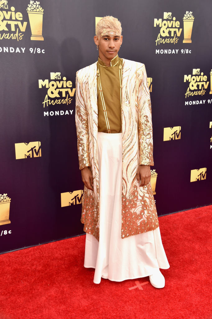 Keiynan Lonsdale at the 2018 MTV Awards red carpet; he is wearing a cream-coloured skirt, a camel-coloured top and a embroidered jacket of similar colours 