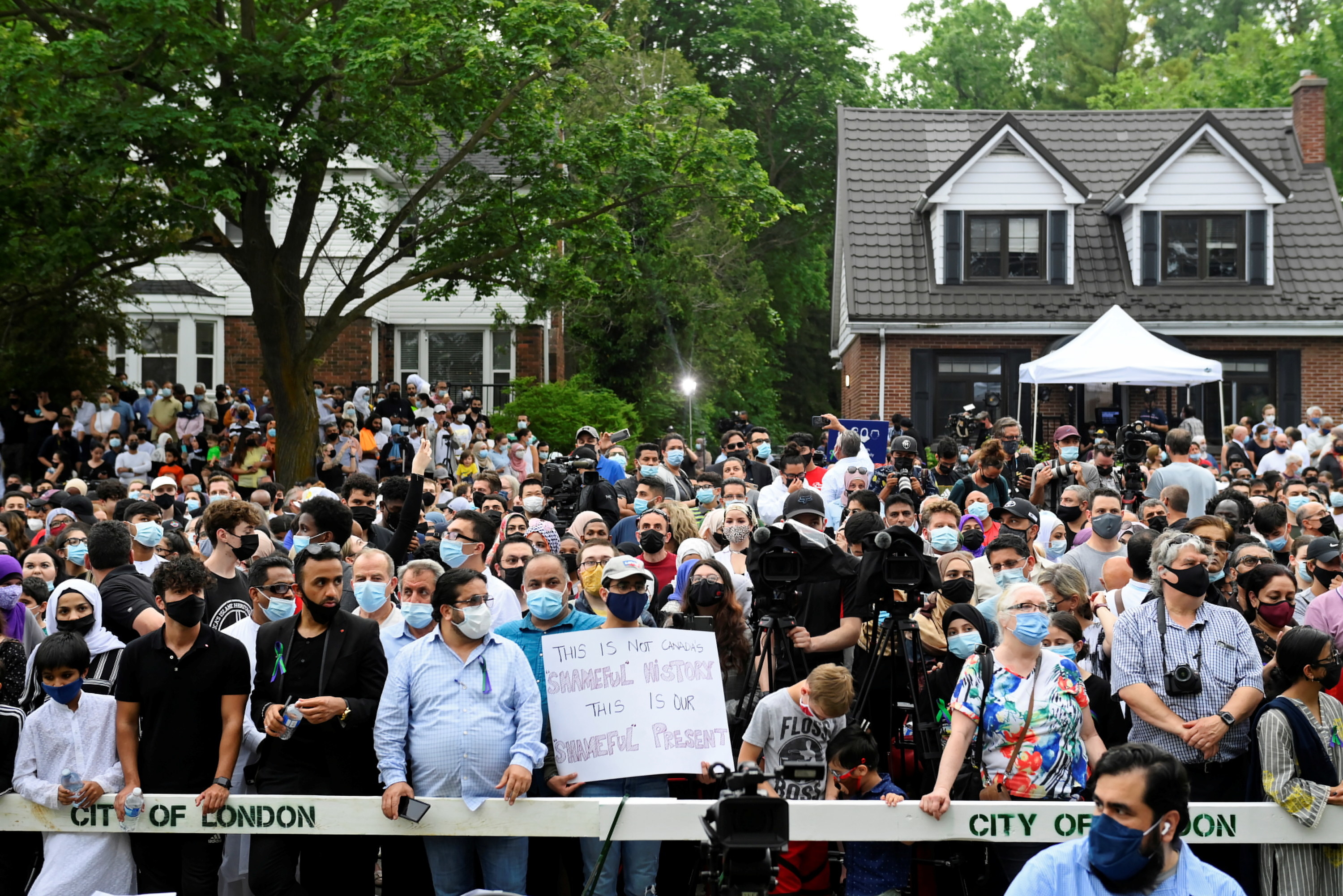 A crowd of people, most of them masked, stand outside behind a &quot;City of London&quot; barrier.