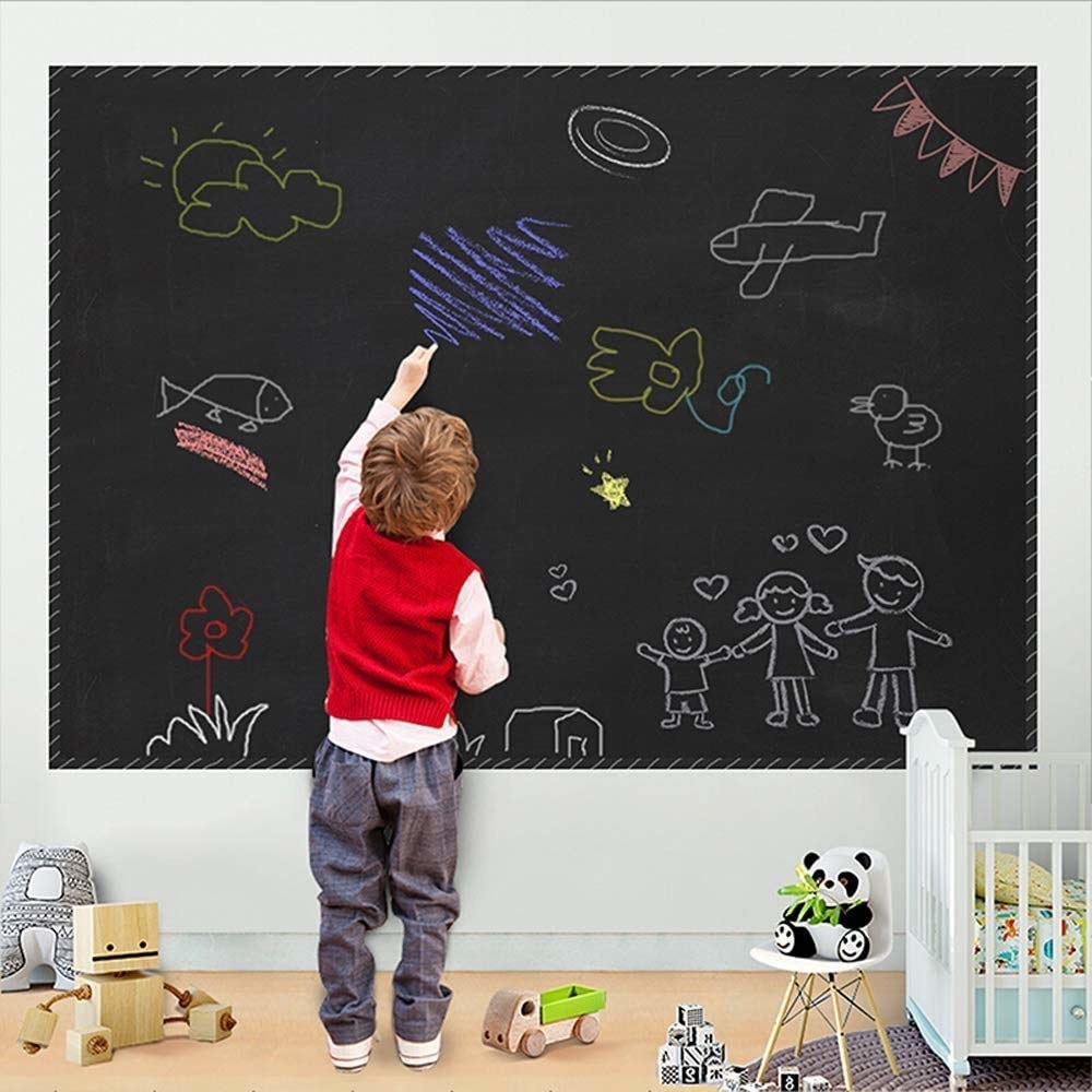 A child scribbling on a paper blackboard with chalk that is stuck to a wall.