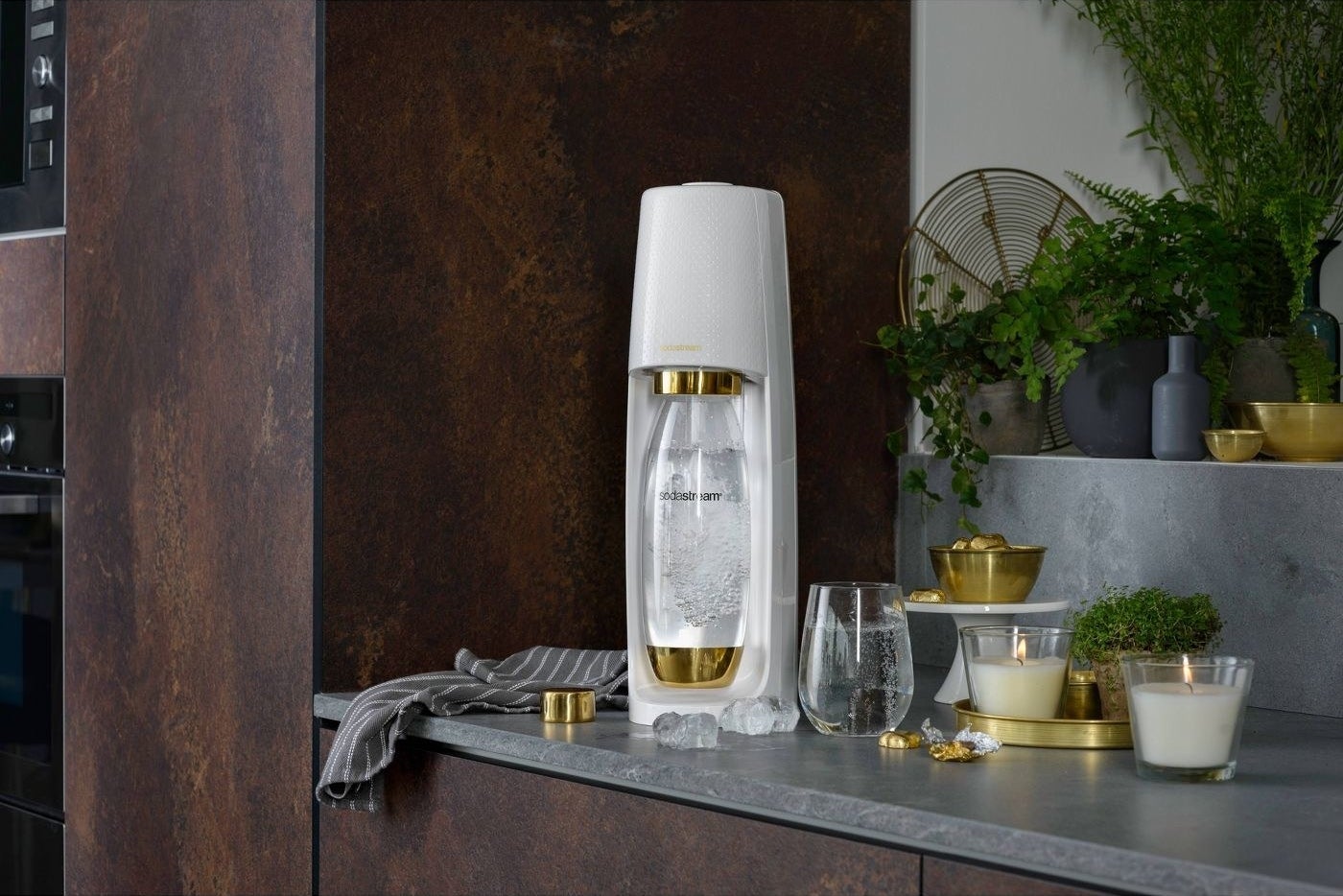 white and gold sodastream with sparkling water inside next to a glass and candles