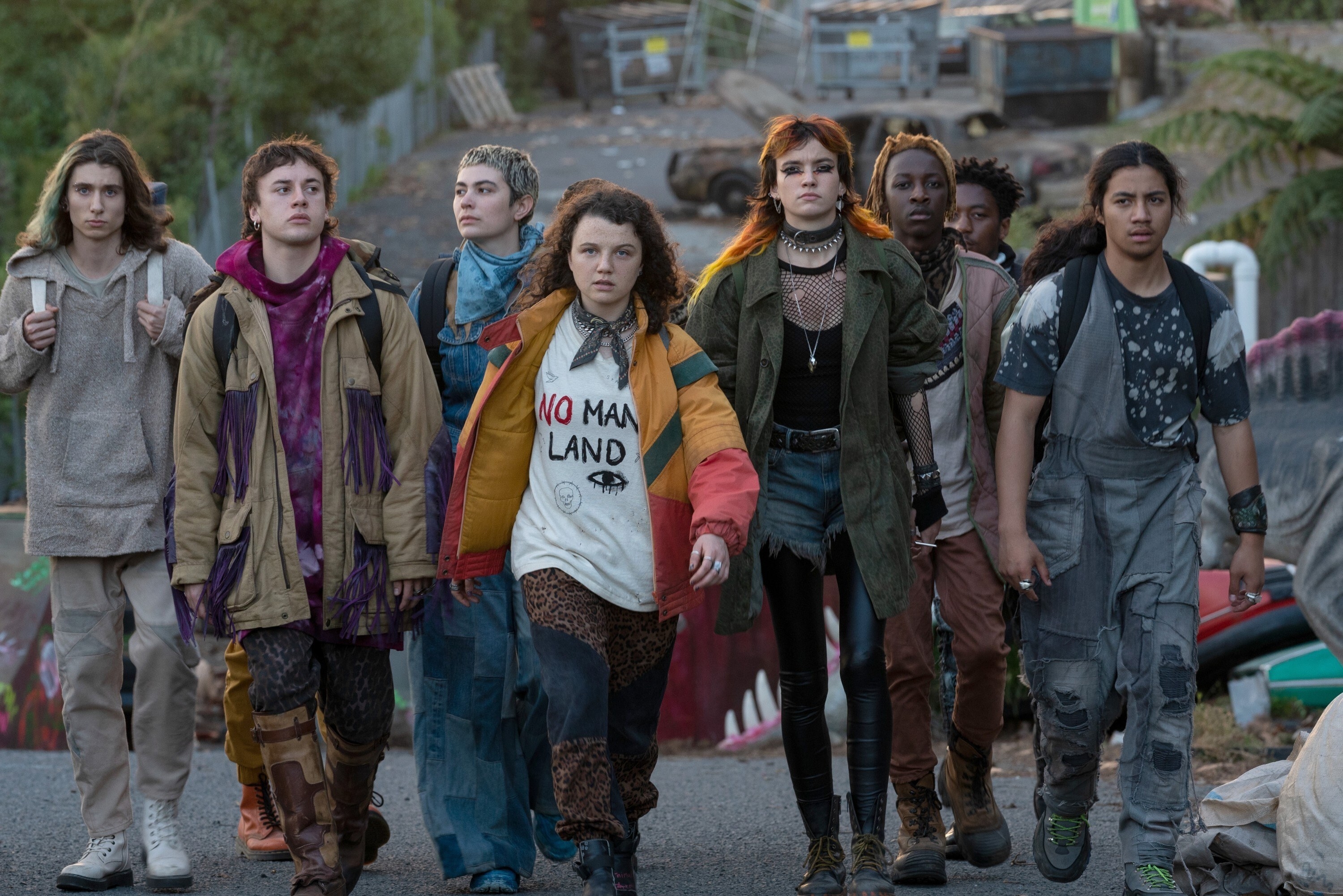 Bear in front of a group of young people dressed in somewhat punk outfits, looking sullen and walking together across a landscape with a burned-out car in the background