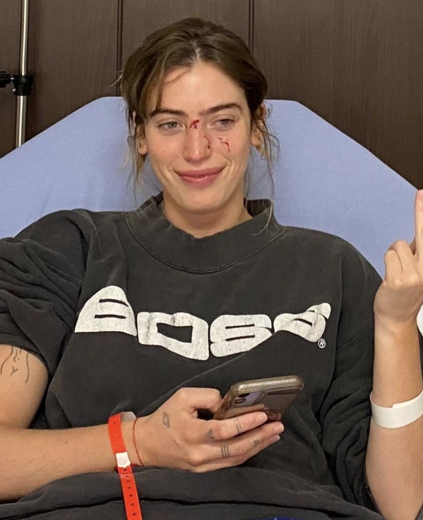 Clara flips off the camera while sitting in a hospital bed in a black sweatshirt