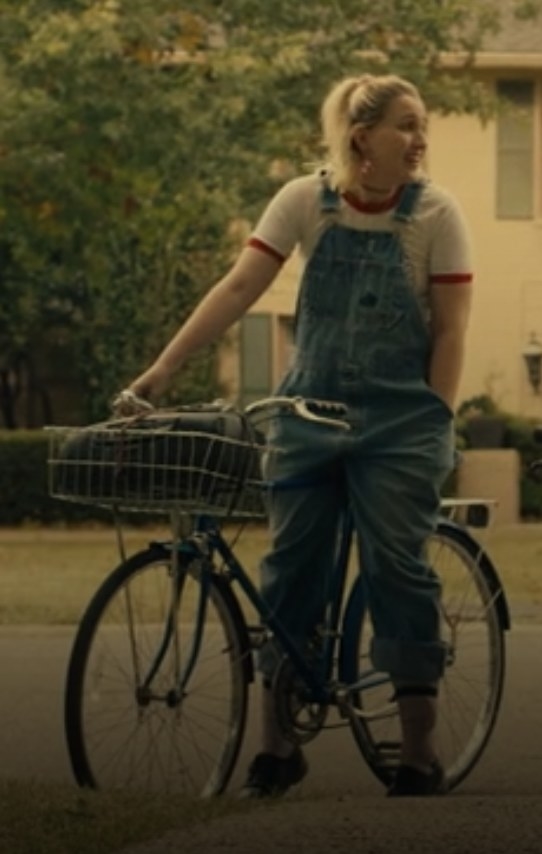 A blond girl sitting on a bike in denim overalls with cuffed bottoms.