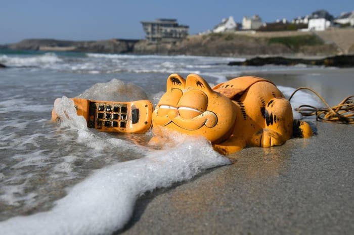 A phone shaped like Garfield the cat sitting in the tides and sea foam on a beach