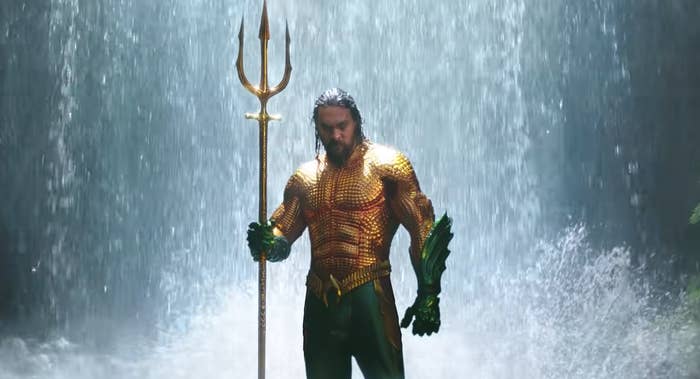 Aquaman stands in the middle of a waterfall while wearing his iconic gold and green costume while holding a giant triton. 