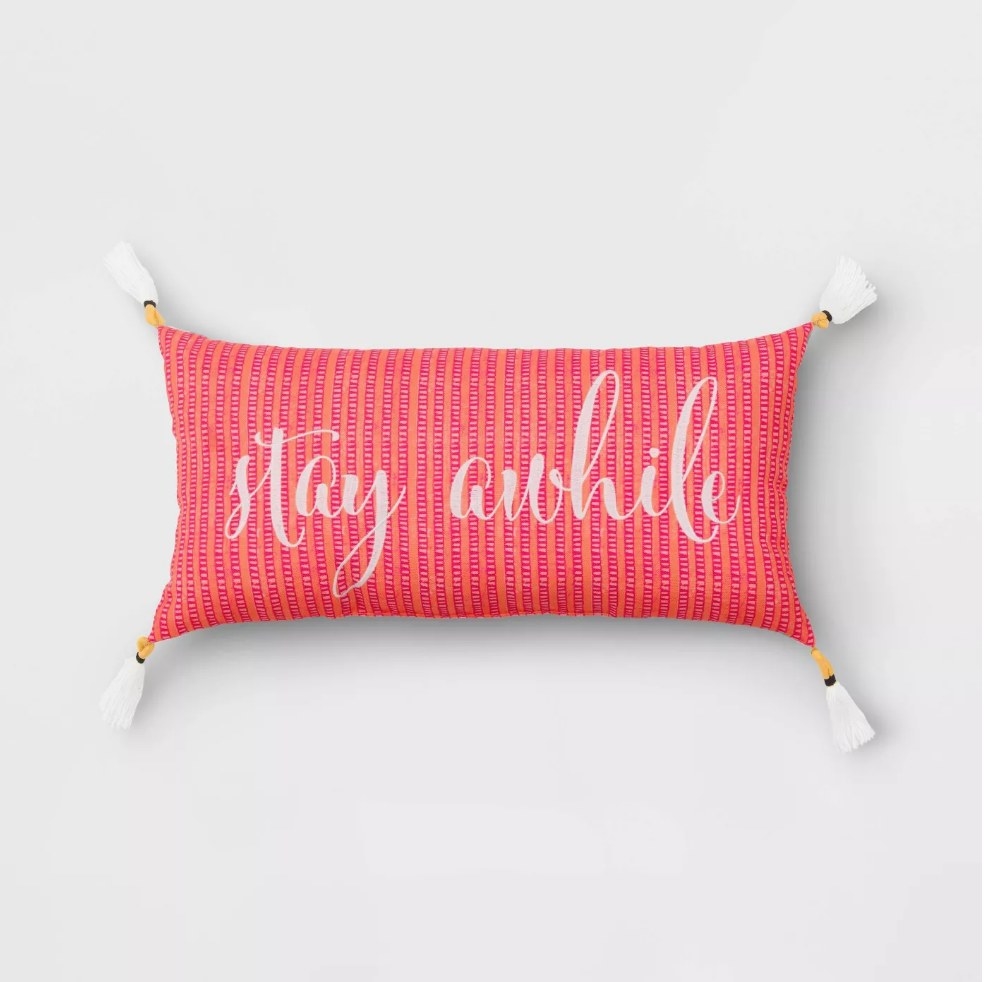 Pink and orange outdoor lumbar pillow with greeting &quot;Stay Awhile&quot; and white tassels on the ends 
