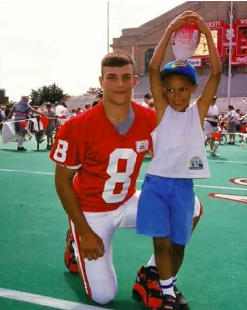 small kid posing with his arms around his head next to a football player