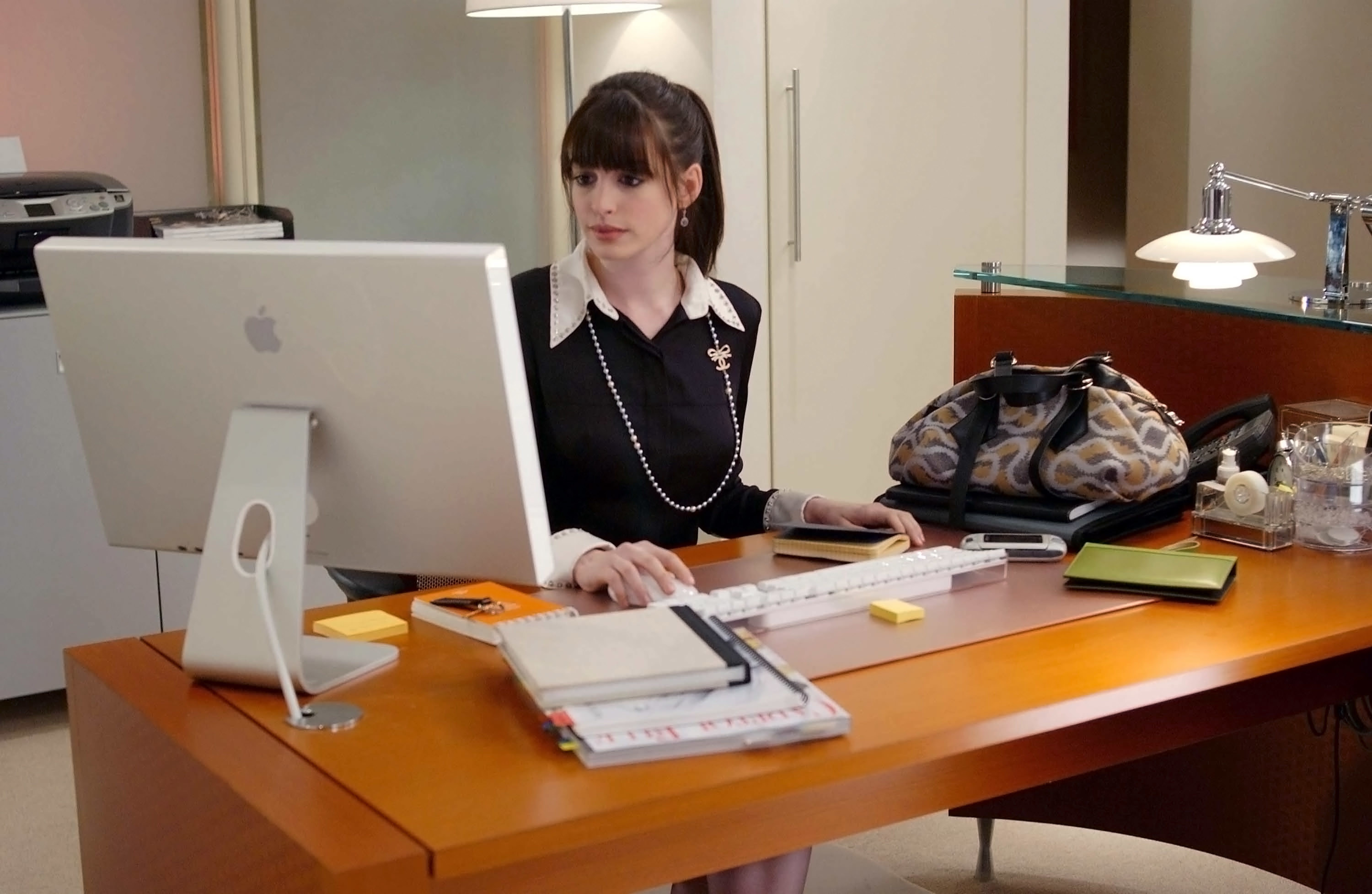 Anne sits at a desk in a scene from Devil Wears Prada