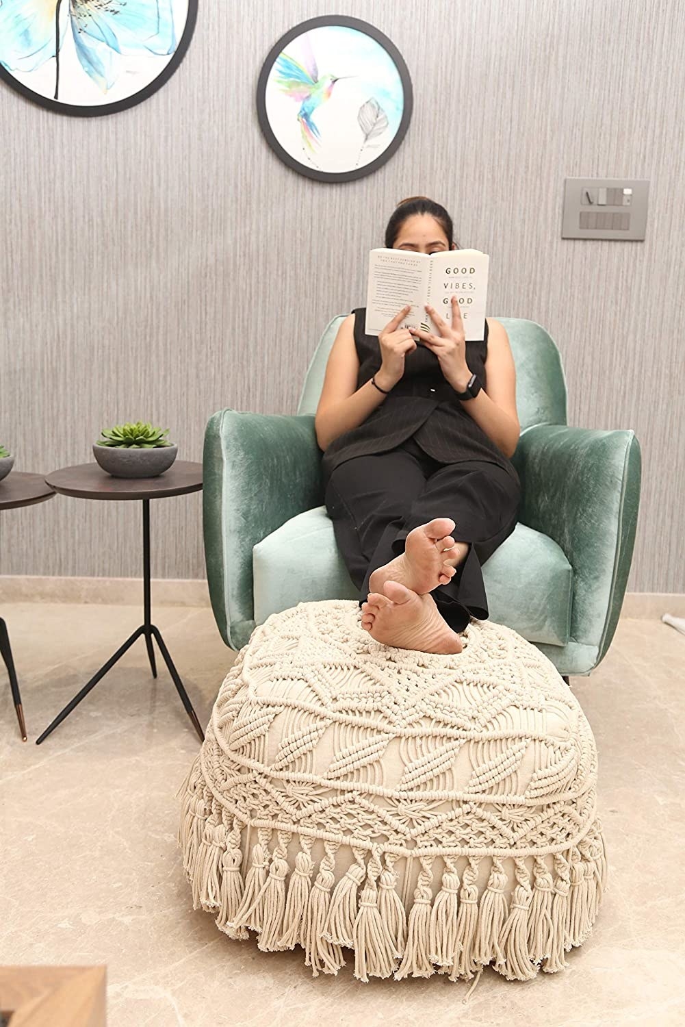 A woman reading with her feet placed on a beige macrame ottoman. 
