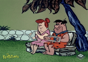 Fred Flintstones sitting on a rock bench helps his wife wind yarn into a ball. 