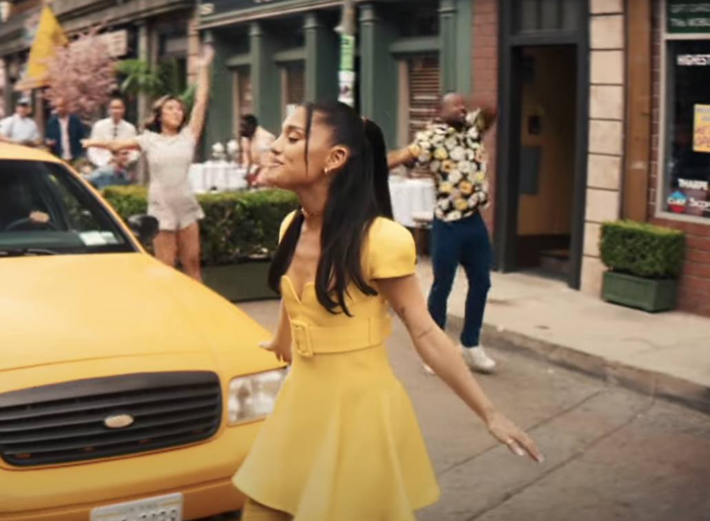 Ariana Grande wears a yellow dress in front of a yellow taxi cab 