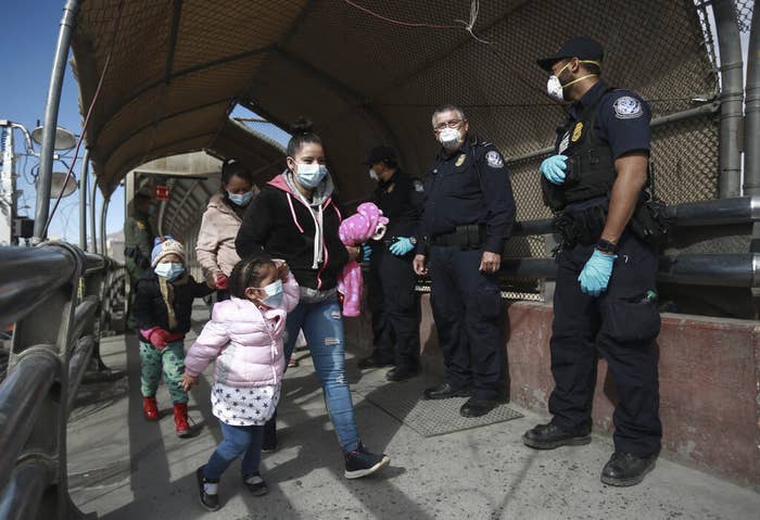 Two young children holding hands with adults walk past border authorities